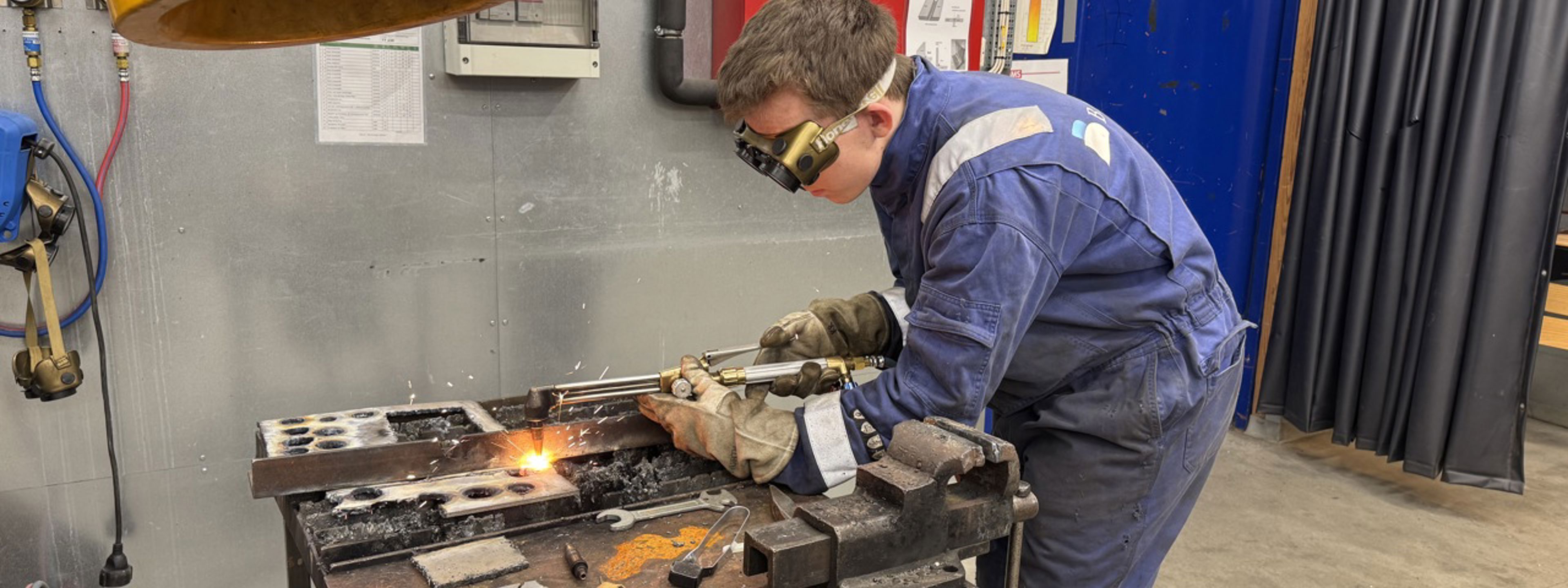 Gut i arbeidsdress og vernebriller står bøyd over ein arbeidsbenk og sveiser. Det står ein liten ildsprut ut frå det han arbeider med