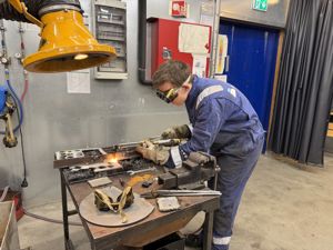 Gut i arbeidsdress og vernebriller står bøyd over ein arbeidsbenk og sveiser. Det står ein liten ildsprut ut frå det han arbeider med