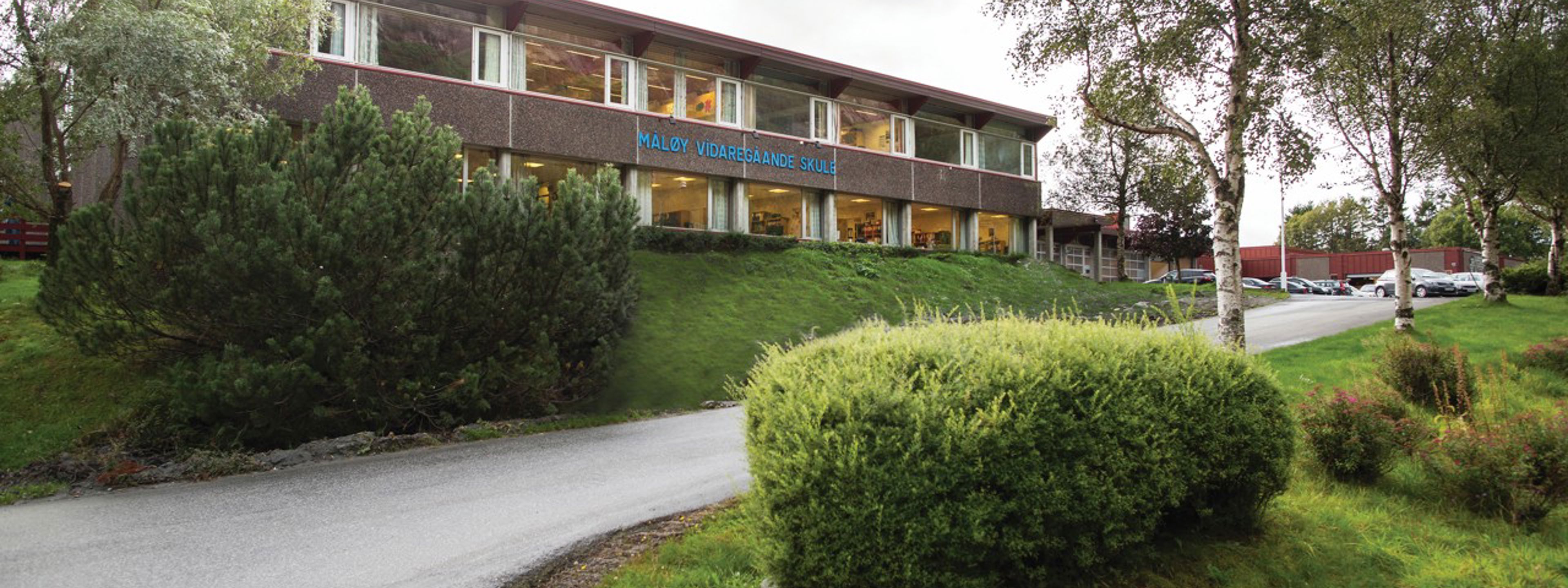 Framsida av skulen sitt hovudbygg. Det står "Måløy vidaregåande skule" med blå bokstavar på veggen.